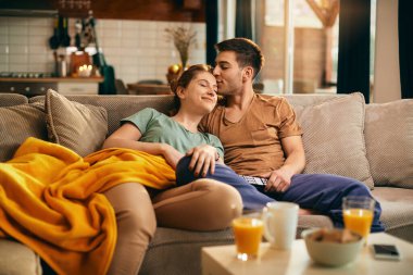 Young loving couple relaxing on the sofa and watching TV. Man is kissing his girlfriend. 