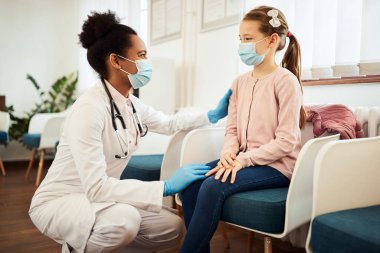 Smiling African American pediatrician communicating with a little girl in waiting room at doctor's office and wearing face mask due to COVID-19 pandemic.