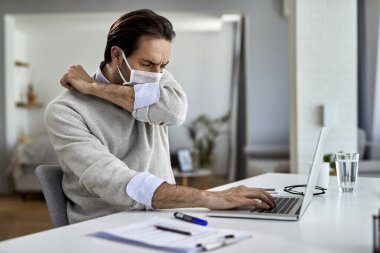 Young businessman coughing into elbow while wearing protective face mask and working on a computer at home. 