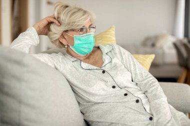Senior woman thinking of something and wearing face mask while relaxing at home during COVID-19 pandemic.