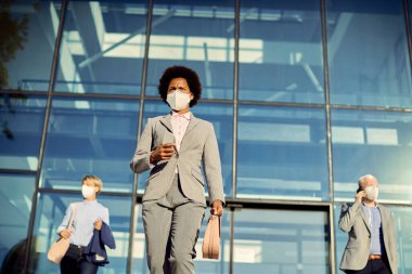Low angle view of African American businesswoman with protective face mask leaving office building. Her colleagues are in the background.  