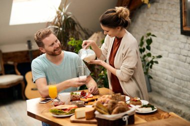 Happy man enjoying while his wife is poring him milk into a glass during breakfast at home.
