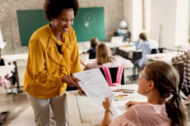 Happy black elementary teacher giving test results to schoolgirl on a class in the classroom. 