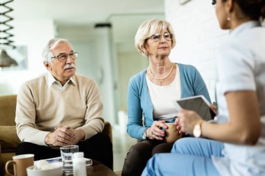 Senior couple talking to healthcare worker during home visit. Focus is on senior woman. 