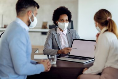 Happy black bank manager using computer while talking to a couple during the meeting in the office. All of them are wearing protective face masks due to coronavirus pandemic. 