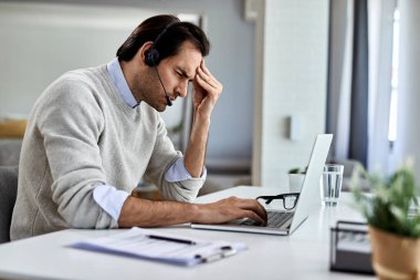 Young businessman holding his head in pain while working on a computer at home. 