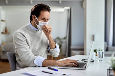 Young entrepreneur coughing while wearing protective face mask and working on a computer at home. 