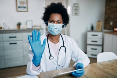 African American doctor working on digital tablet at her office and waving while looking at camera. She is wearing protective face mask and gloves due to coronavirus pandemic.