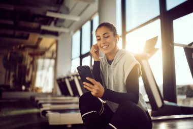 Happy sportswoman using mobile phone while relaxing after sports training in a gym. Copy space.