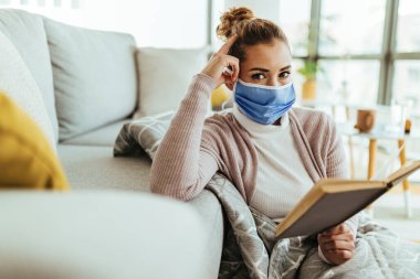 Young woman wearing protective face mask relaxing in her living room and reading a book. 