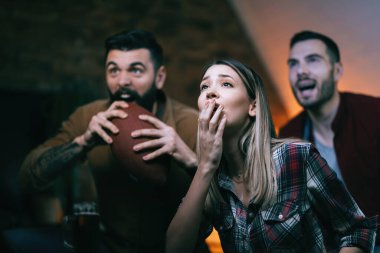 Group of sports fans feeling disappointed while watching rugby match on TV at home. Focus is on shocked woman in the foreground. 