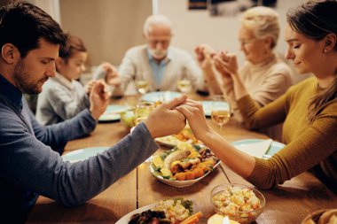 Multi-generation family saying grace while holding hands with eyes closed at dining table. Focus is on foreground.