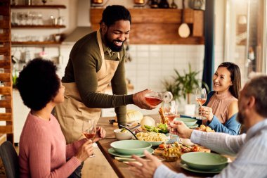 Happy black man pouring wine while having meal with his friends at dining table at home. 
