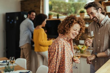 Young couple having fun and laughing while drinking wine in the kitchen. Their friends are preparing food in the background. 