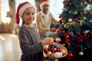 Happy daughter and father decorating Christmas tree together at home. Focus is on daughter looking at camera.