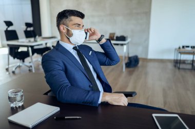 Businessman with face mask talking on smart phone while working in the office during coronavirus epidemic. 