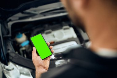 Close-up of mechanic using mobile phone while examining car engine at auto repair shop. Focus is on green touchscreen. Copy space. 