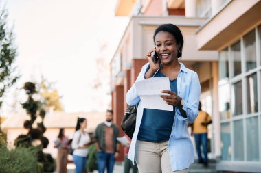Happy African American university student reading test results while communicating over mobile phone outdoors.
