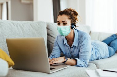 Woman wearing face mask and surfing the net on laptop while relaxing in the living room.