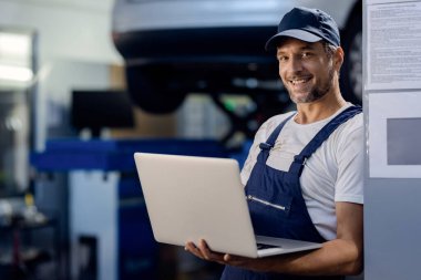 Happy repairman using laptop while working at car workshop and looking at camera. 