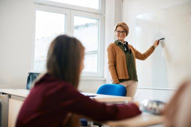 Happy math teacher giving lecture on whiteboard to elementary students in the classroom.