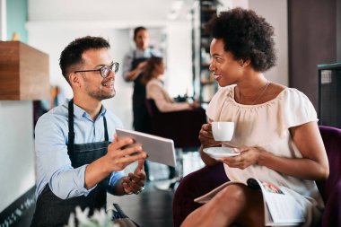 Happy black woman drinking coffee and talking to her hairstylist who is showing her something on touchpad at hair salon.
