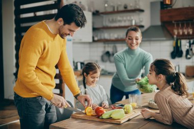 Happy parents with two daughters preparing healthy food in the kitchen. Focus is on man. 