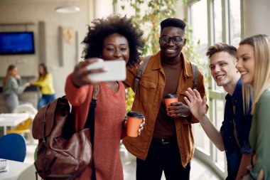 Group of happy university friends having fun while taking selfie with smart phone during coffee break at cafeteria. 