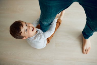 High angle view of carefree kid having fun while holding on to his fathers leg at home and looking at camera.  