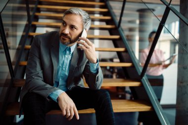 Businessman using mobile phone and talking to someone while relaxing on steps indoors.