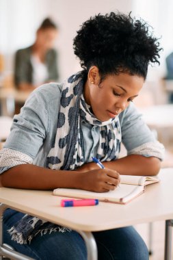 Mid adult black woman writing in notebook while having lecture at university classroom.