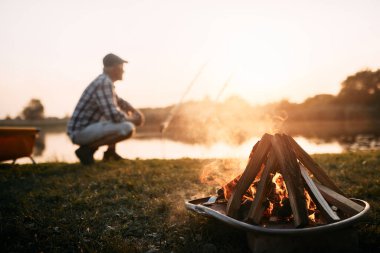 Close-up of bonfire with man fishing on the river in the background at sunset. Copy space.