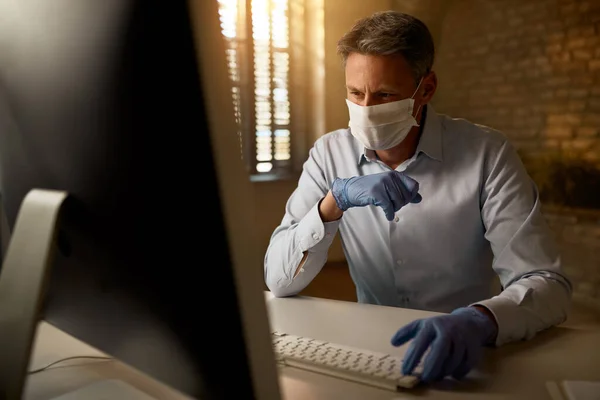 Pensive businessman wearing face mask while working on desktop PC in the office during virus epidemic.