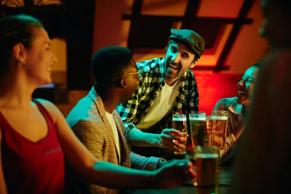 Young happy man and his friends having fun and toasting with beer while gathering in a pub at night.