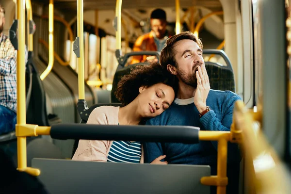 Tired man yawning while his black girlfriend is napping on his shoulder in a city bus.