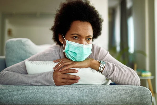 Distraught African American woman with face mask lying down on sofa and thinking while being at home during virus pandemic.