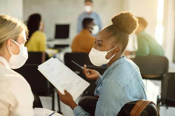 African American college student with a face mask talking to her friend about exam results in lecture hall.