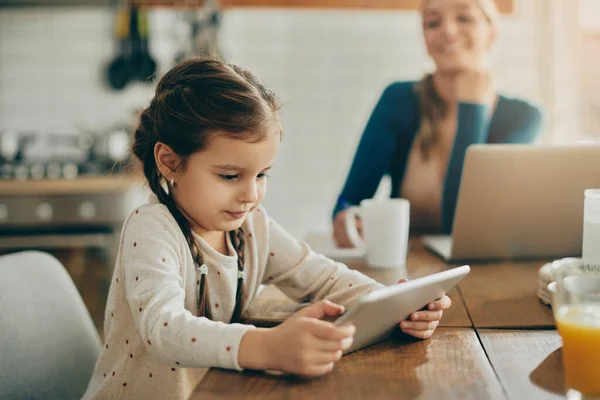Little girl using touchpad and watching something on the Internet while her mother is working on computer in the background.