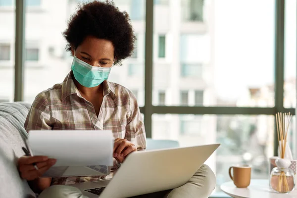 Black woman with face mask reading business reports while working on laptop at home during virus pandemic.