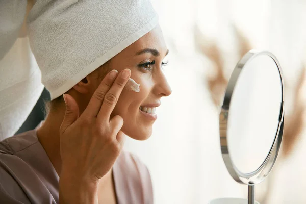 Young smiling woman using moisturizer while taking care of her face skin at home.
