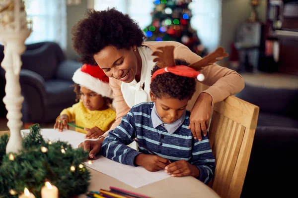 Happy African American mother enjoying with her children who are drawing on paper during Christmas day at home.