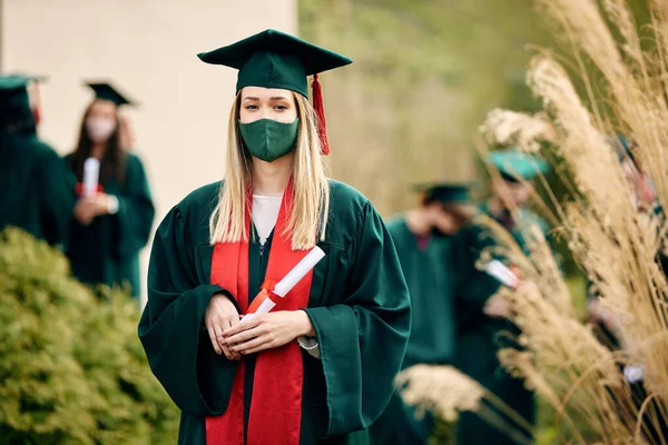Portrait of female graduate with university certificate wearing face mask and looking at camera.