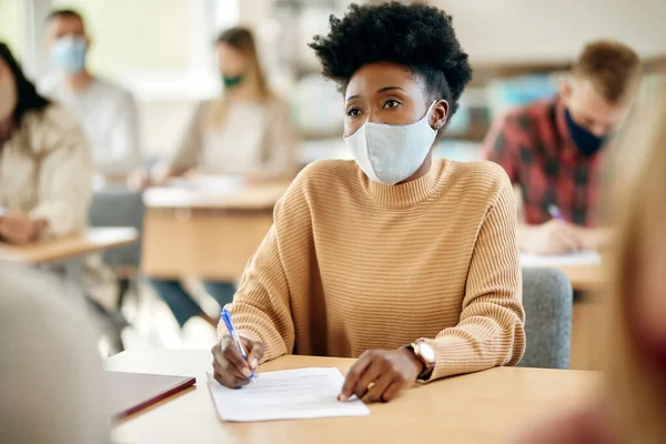 African American college student taking notes while wearing face mask in the classroom during coronavirus pandemic.
