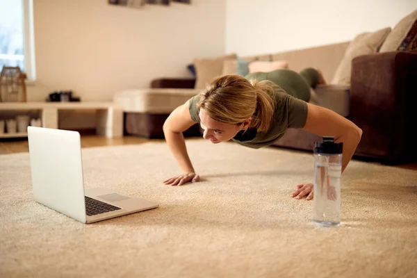 Athletic woman following online exercise class on laptop while doing push-ups at home.