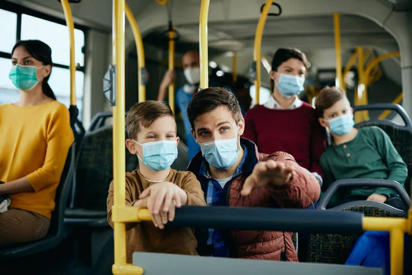 Happy father and son with face masks traveling by public bus. father is pointing at something in the distance.