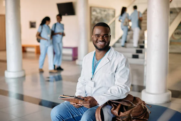Happy African American medical student in white lab coat sitting in corridor at medical university and looking at camera.