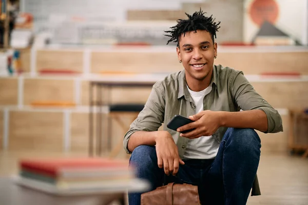 Portrait of young happy black student using mobile phone at university library looking at camera.