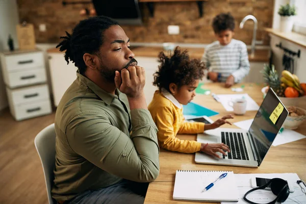 Distraught black working father thinking of something while daughter is sitting on his laptop and using his laptop at home.