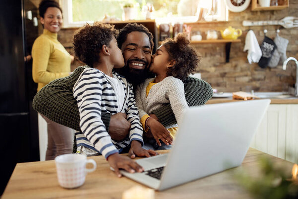 Happy black man being kissed by his children while using laptop at home. Mother is in the background. 
