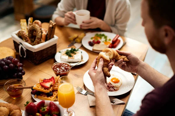 Close-up of man eating croissant while having breakfast with his girlfriend at dining table.